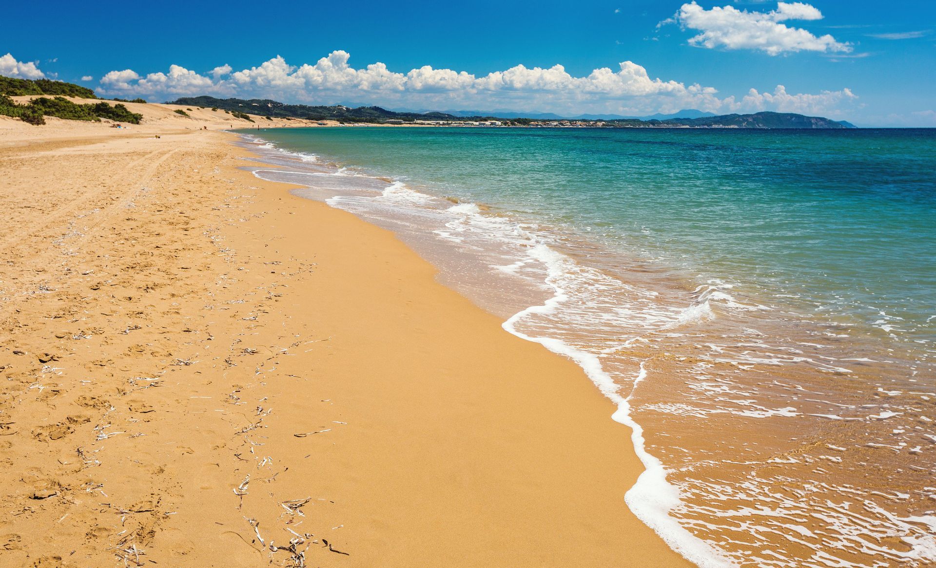 Une large plage de sable doré s'incurve le long de l'océan turquoise calme sous un ciel bleu parsemé de nuages blancs moelleux.