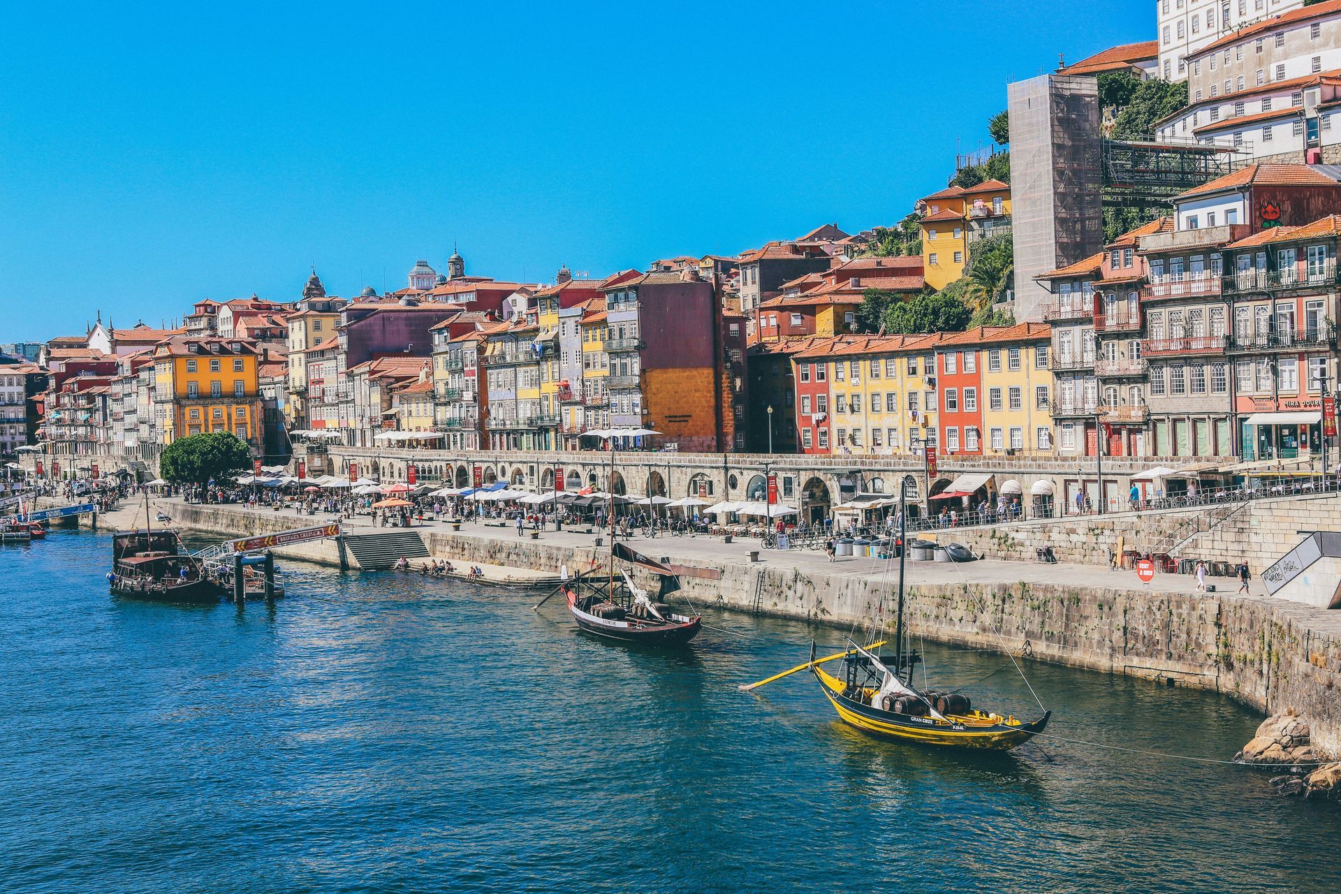 Traditional boats float on a river next to a bustling waterfront lined with colorful, historic buildings under a clear blue sky.