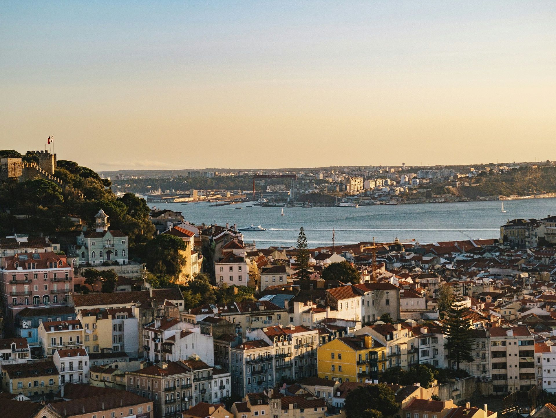 A cityscape of colorful buildings and red-tiled roofs overlooks a wide river at sunset, with a historic castle on a hill.