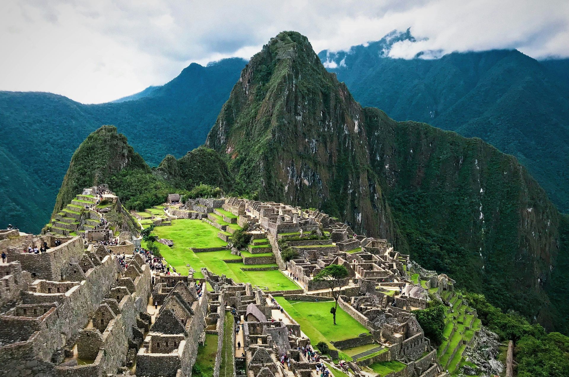 A high-angle view of ancient stone ruins and green terraces nestled in lush mountains, with a prominent peak rising against a cloudy sky.