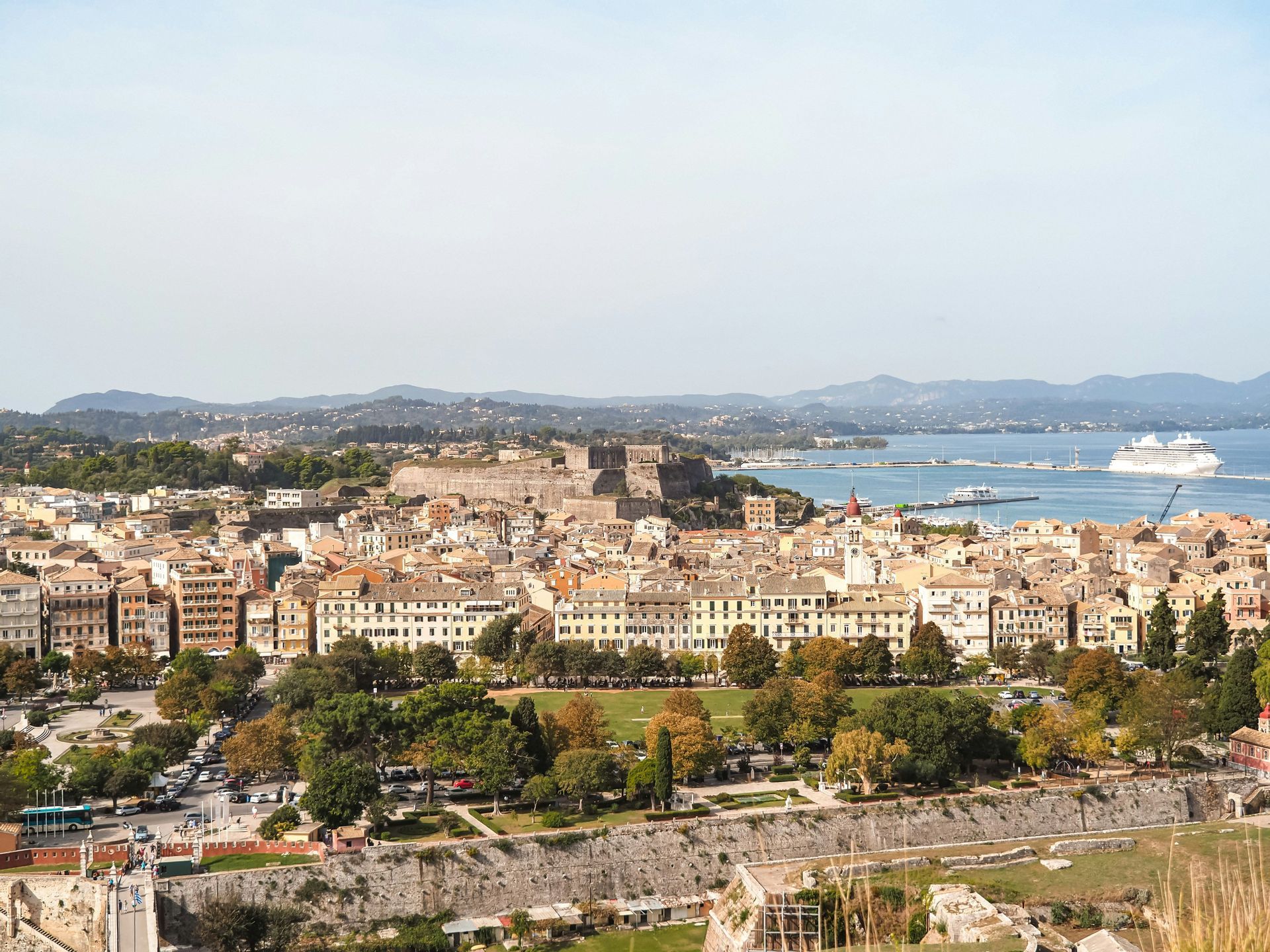 An aerial view of a historic coastal city featuring a fortress, a park, and a large cruise ship in the harbor.