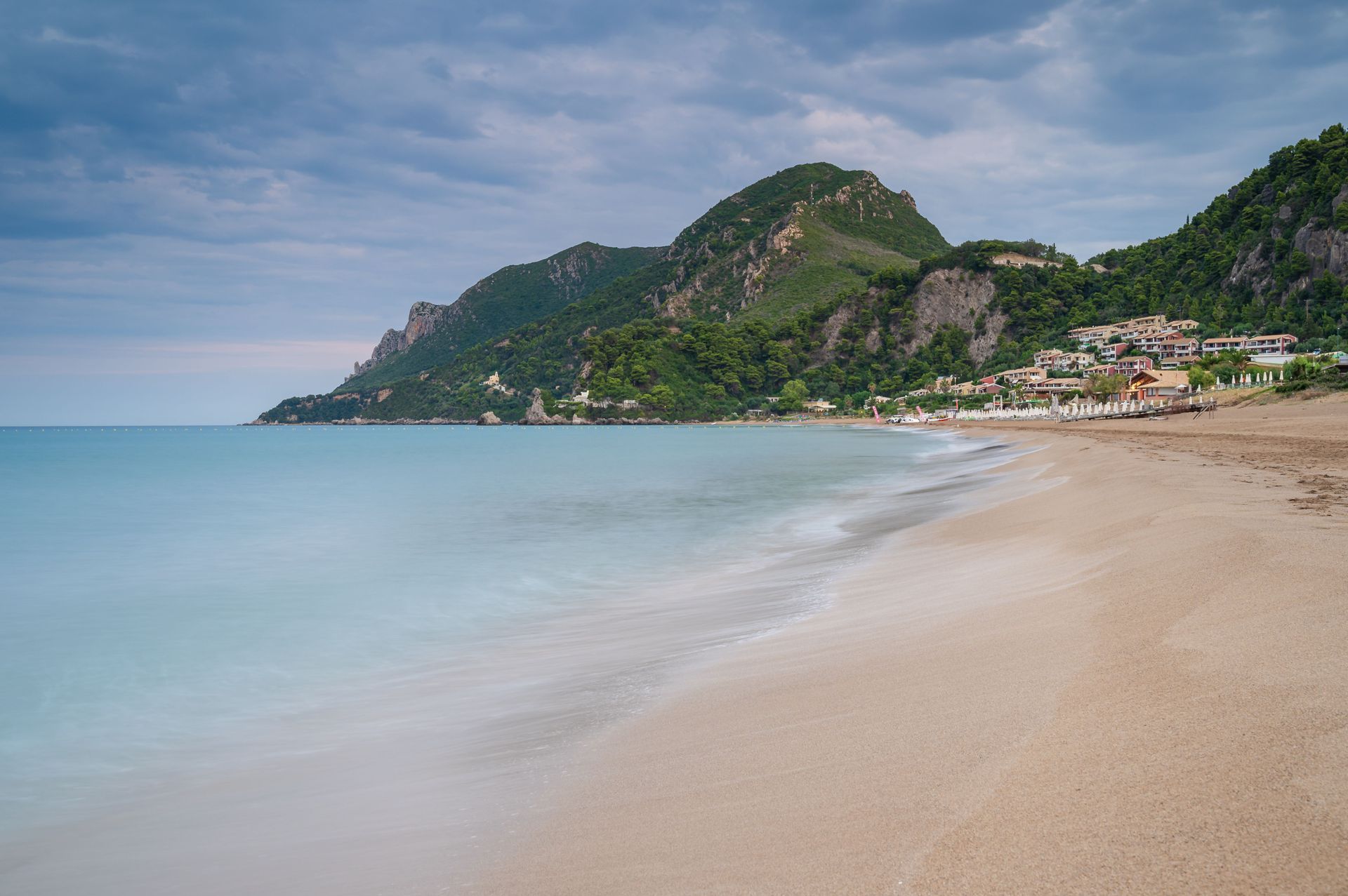 A long-exposure shot of turquoise water washing onto a sandy beach, with a green mountain and coastal buildings in the background.