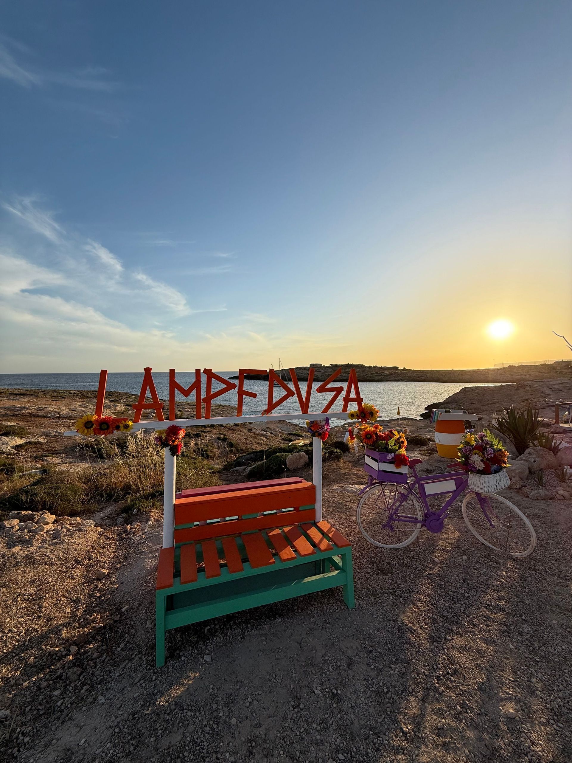 Un grande cartello con lettere arancioni, una panchina colorata e una bicicletta decorativa con fiori su una riva rocciosa al tramonto.