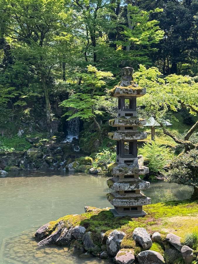 Un'alta lanterna pagoda di pietra si erge su una riva ricoperta di muschio, accanto a uno stagno, in un giardino rigoglioso con una cascata.
