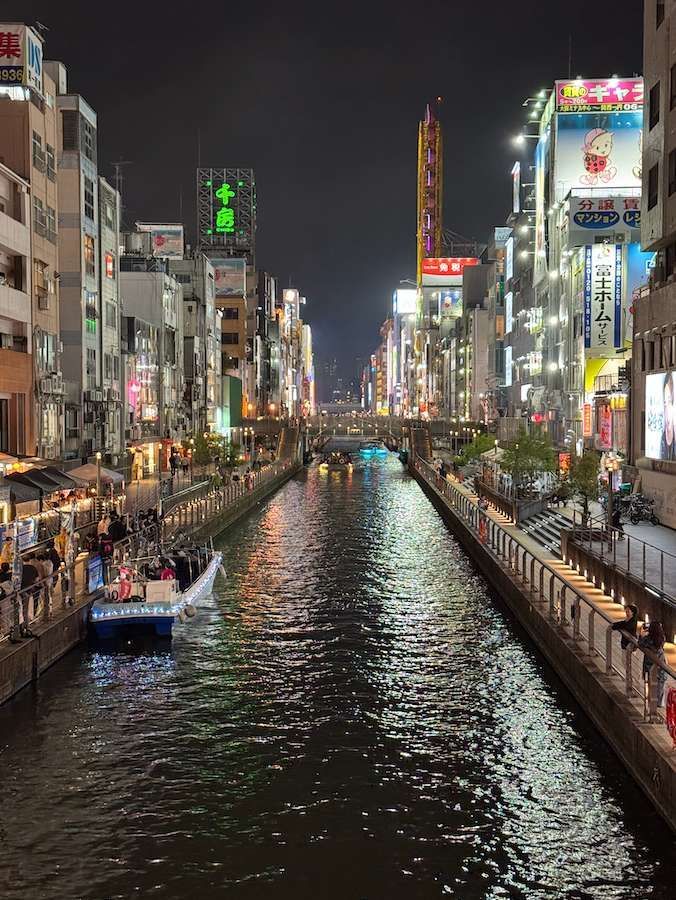 Un canale cittadino di notte, con barche sull'acqua e le sponde fiancheggiate da edifici splendidamente illuminati e insegne al neon.