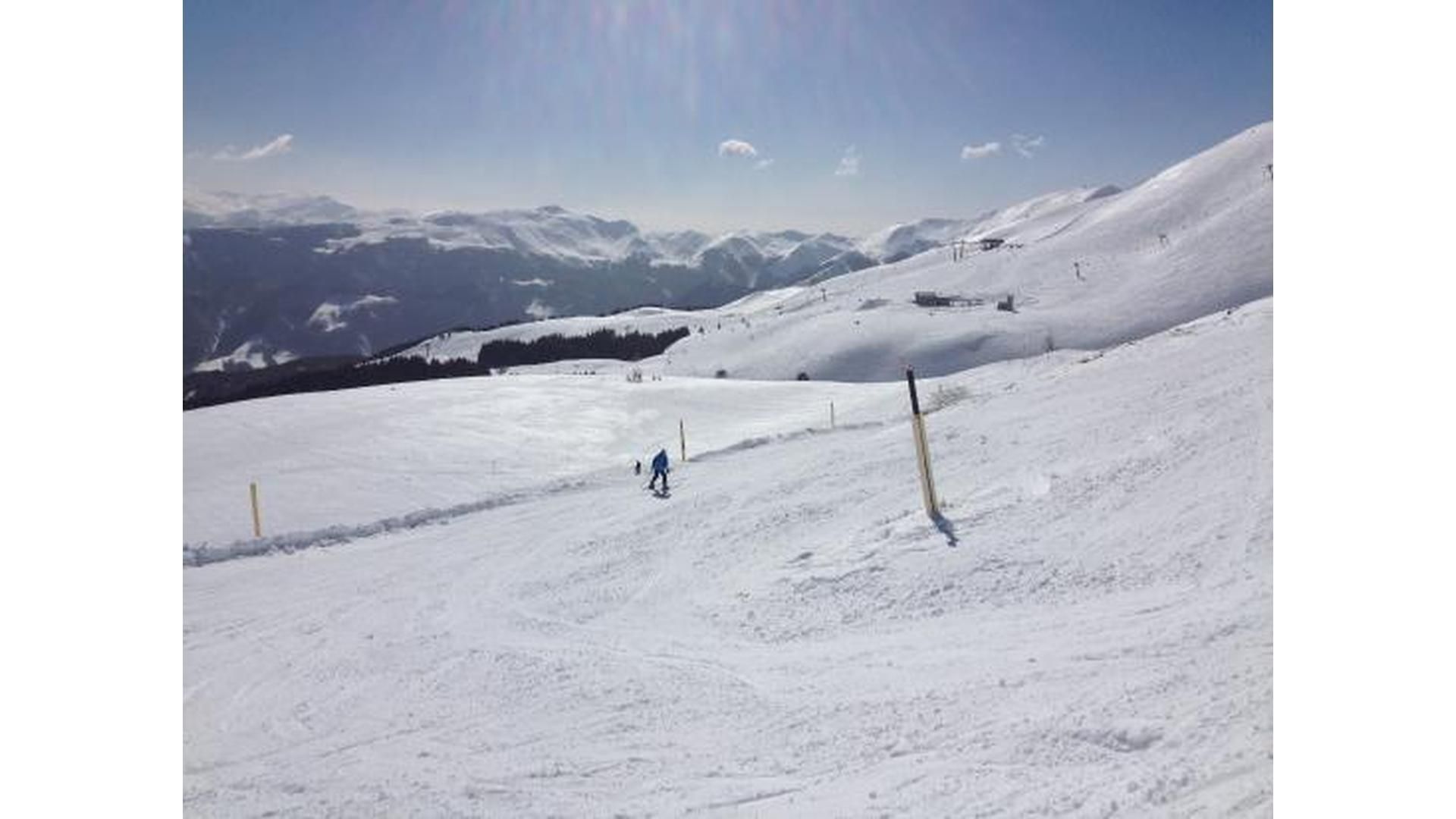 Uno sciatore in giacca blu scende su un vasto pendio innevato, con una catena montuosa in lontananza sotto un cielo soleggiato.