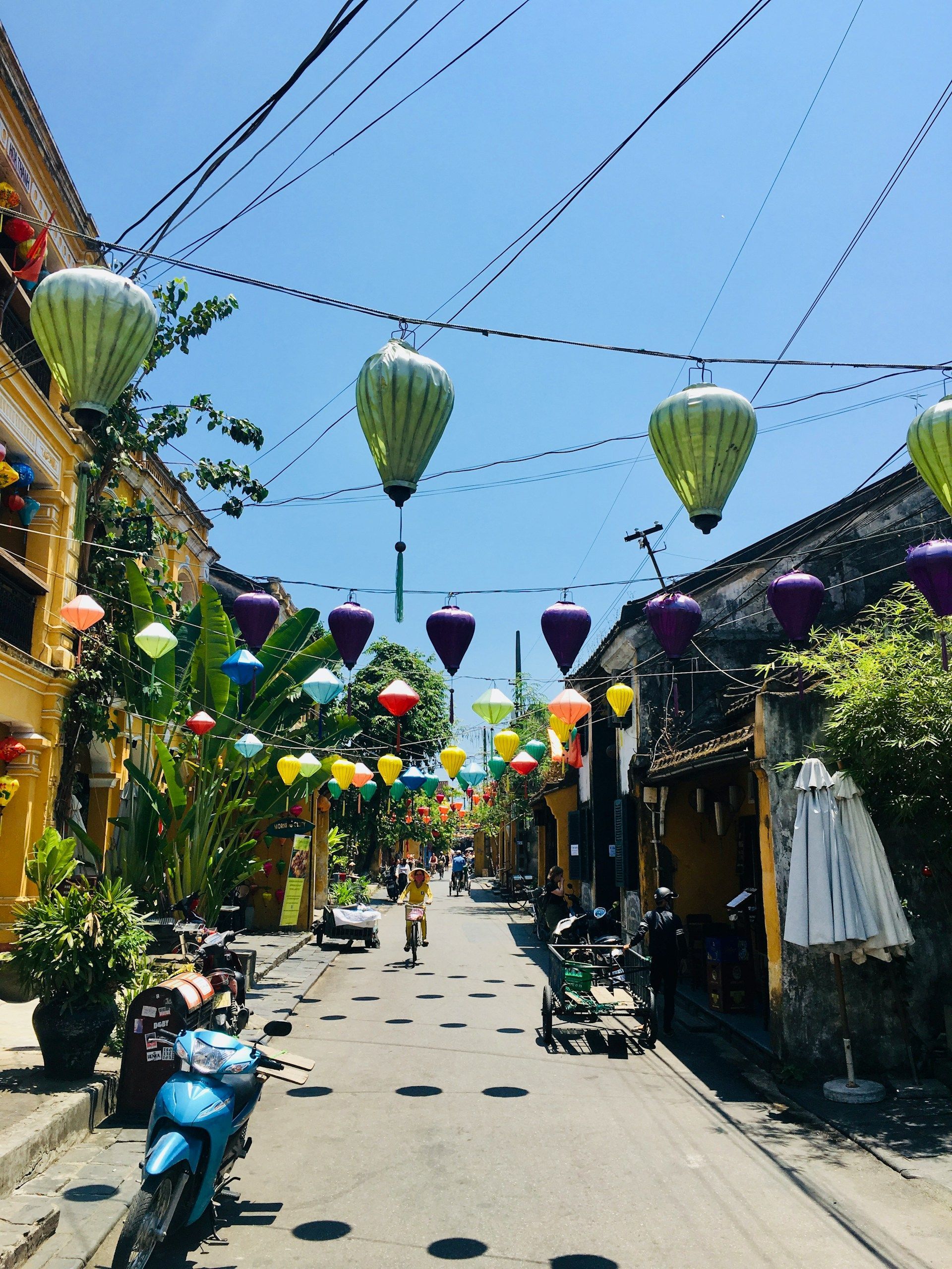 Lanterne colorate adornano una strada stretta e illuminata dal sole, fiancheggiata da edifici gialli e scooter parcheggiati.