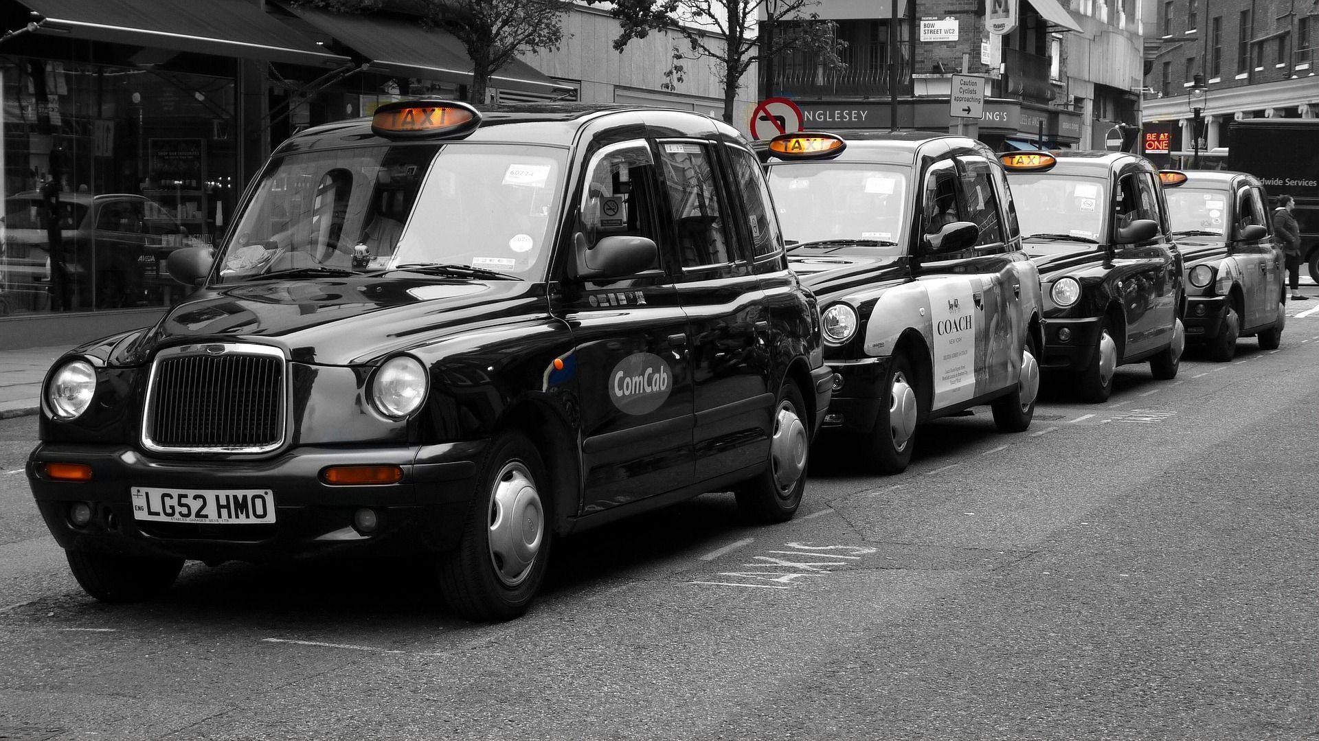 Una fila de taxis negros de Londres estacionados en una calle de la ciudad, capturados en blanco y negro con solo sus letreros de taxi naranjas a color.