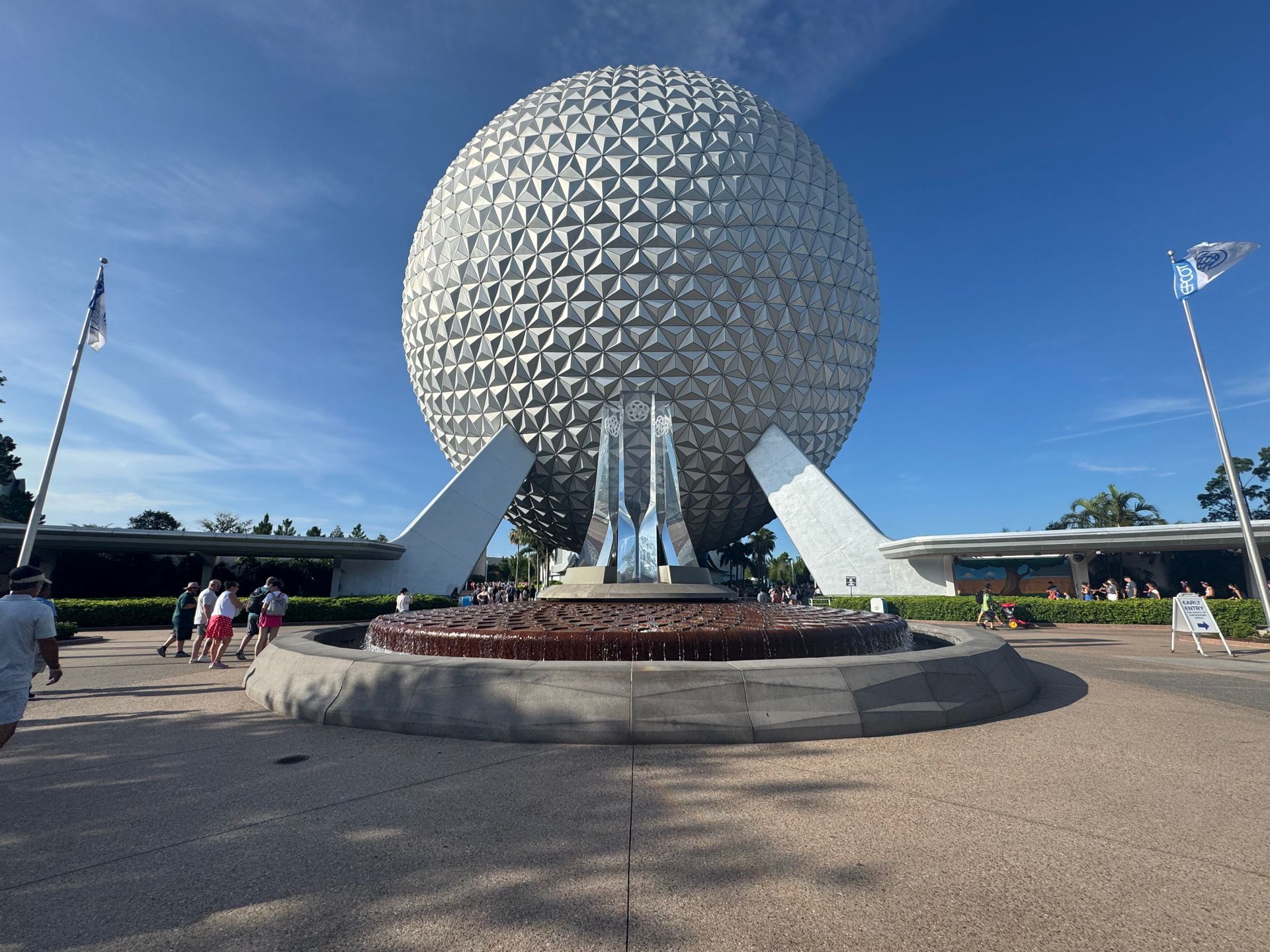 A large geodesic sphere with a triangulated surface stands behind a fountain in a plaza under a clear blue sky.
