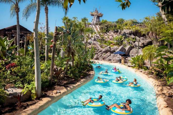 People floating in inflatable rings on a lazy river at a water park, with a large slide and palm trees in the background.