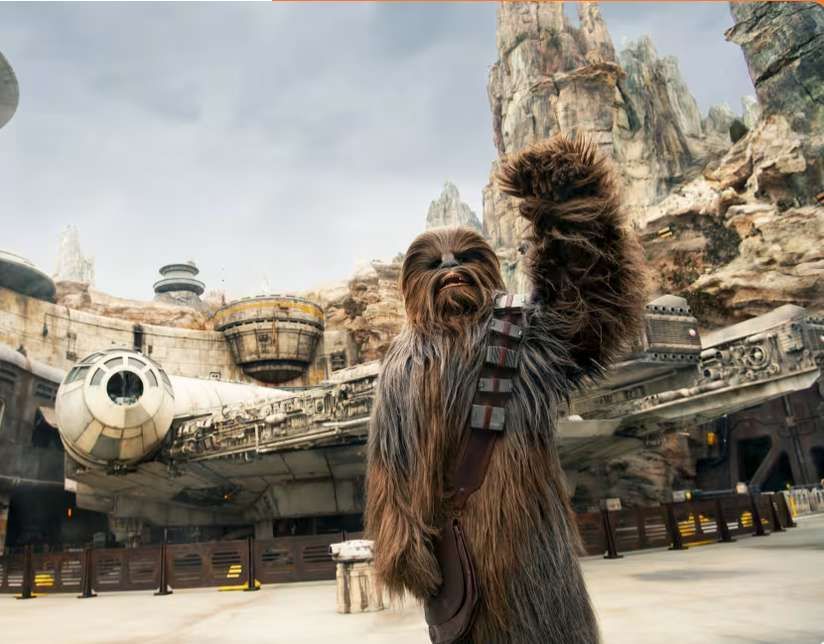 The character Chewbacca raises a furry arm while standing in front of the Millennium Falcon spaceship and rocky cliffs.