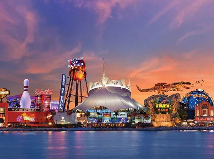 A colorful entertainment complex on the waterfront, featuring a giant white circus tent and themed restaurants, illuminated at sunset.