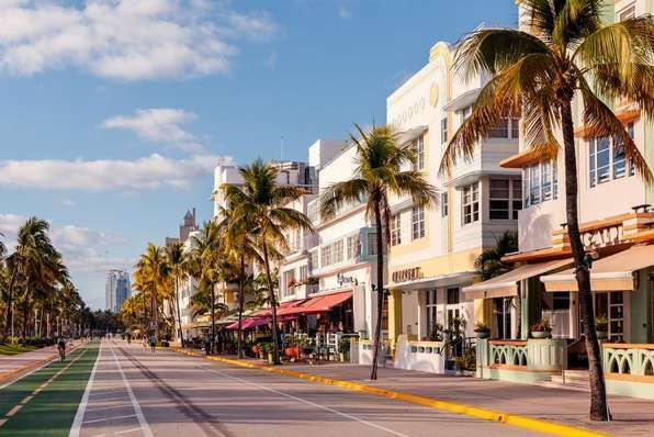 A wide street with a green bike lane is lined with Art Deco buildings and tall palm trees under a sunny, blue sky.