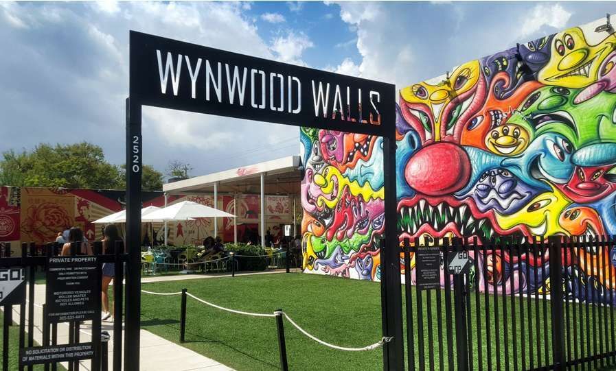 The entrance sign for Wynwood Walls stands next to a building covered in a colorful cartoon-style mural under a blue sky.