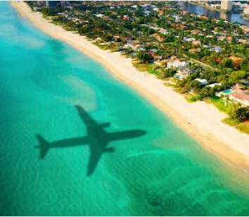 An airplane's shadow is cast over the turquoise ocean water along a sandy coastline.