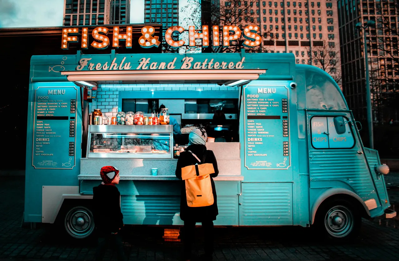 Una mujer y un niño están en el mostrador de un food truck turquesa de fish and chips en un entorno urbano.