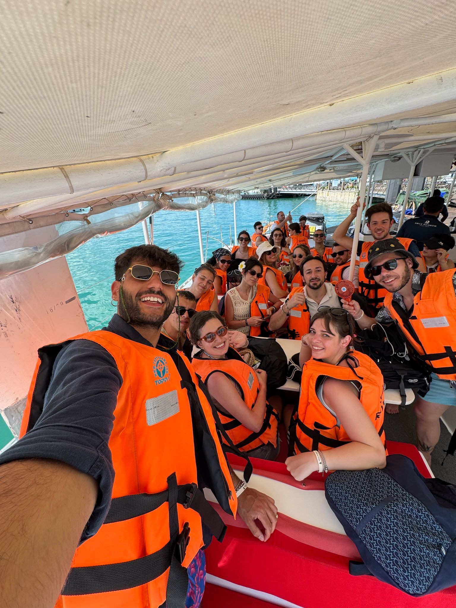 Eine WeRoad-Gruppe macht ein Selfie auf einem Boot, alle in orangefarbenen Schwimmwesten über türkisfarbenem Wasser.
