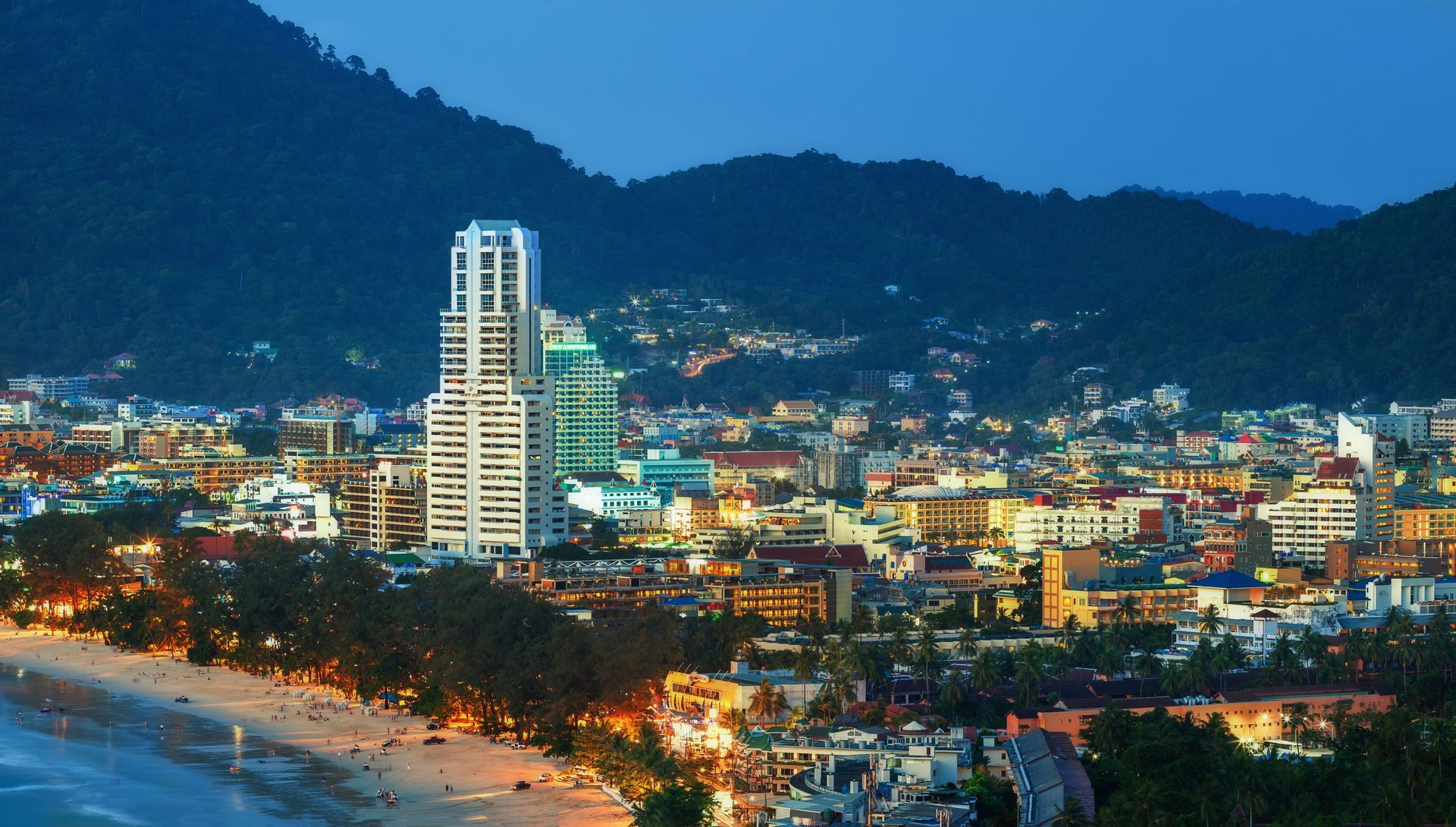 Una ciudad costera iluminada con edificios modernos, enclavada entre una playa de arena y exuberantes colinas verdes al anochecer.