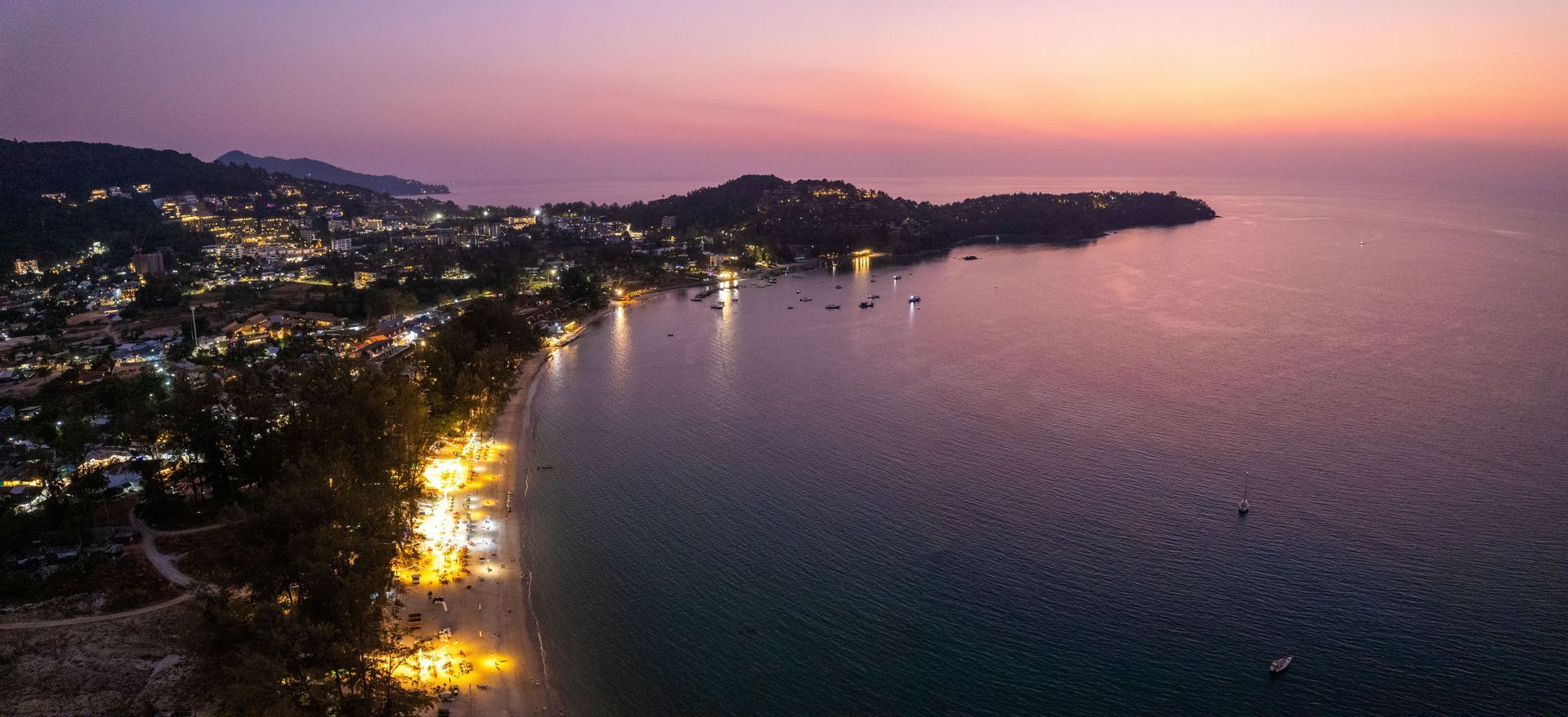 Una vista aérea de un pueblo costero y playa iluminados junto a una bahía tranquila con barcos al atardecer.