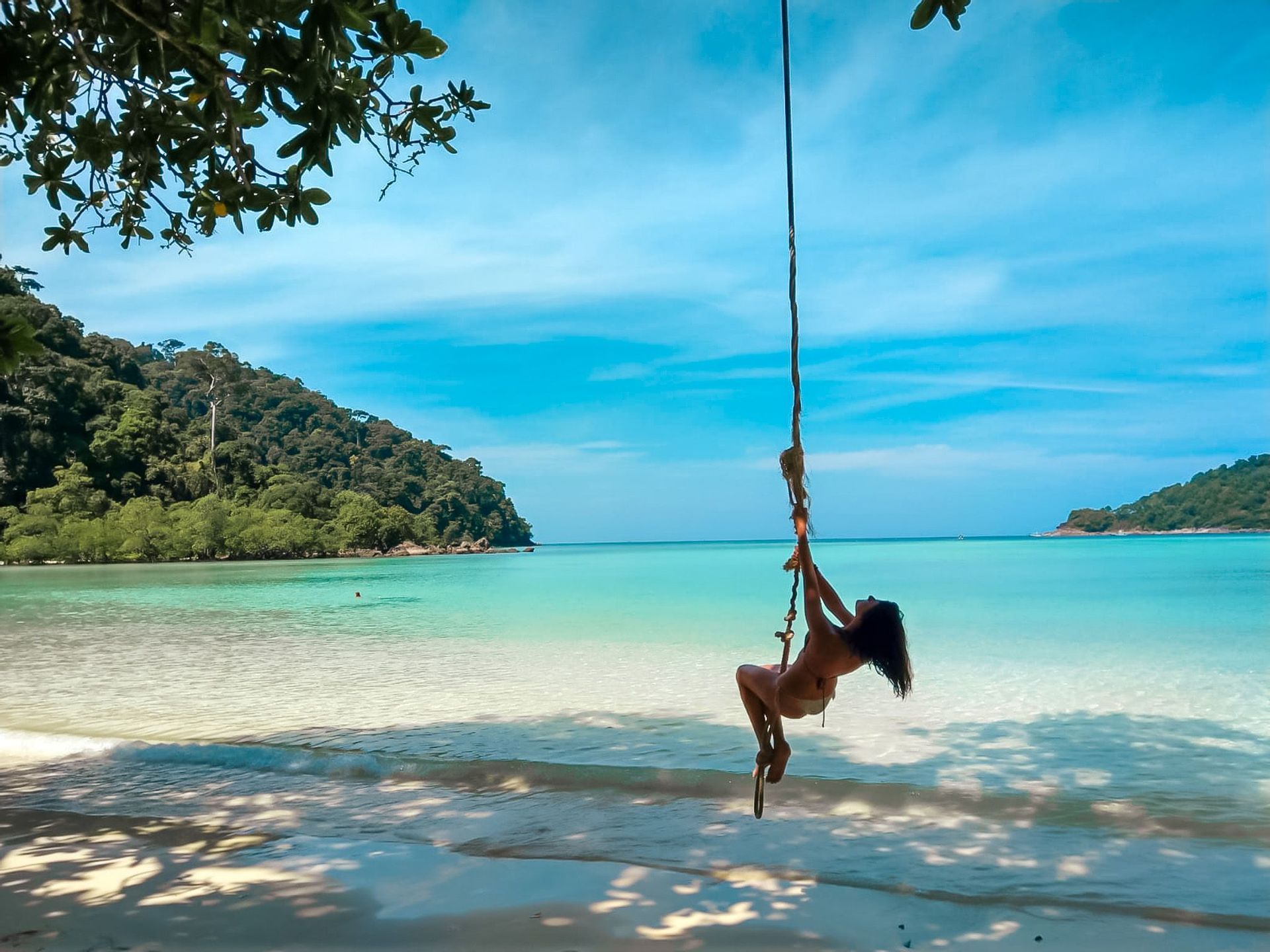 Une femme en bikini se balance sur une corde au-dessus de la mer turquoise depuis une plage de sable blanc avec des collines luxuriantes en arrière-plan.