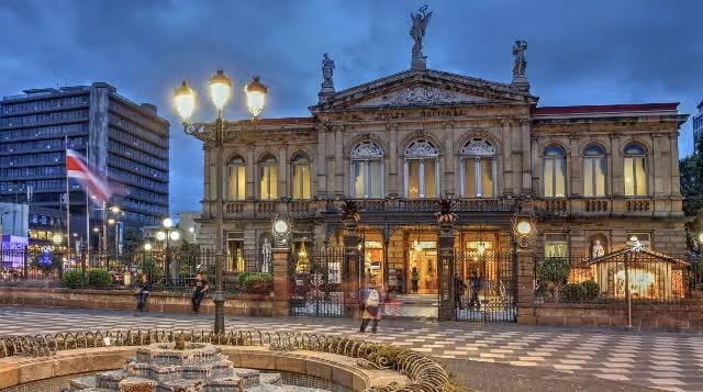 Un sontuoso teatro neoclassico è illuminato su una piazza cittadina al crepuscolo, con una fontana in primo piano.
