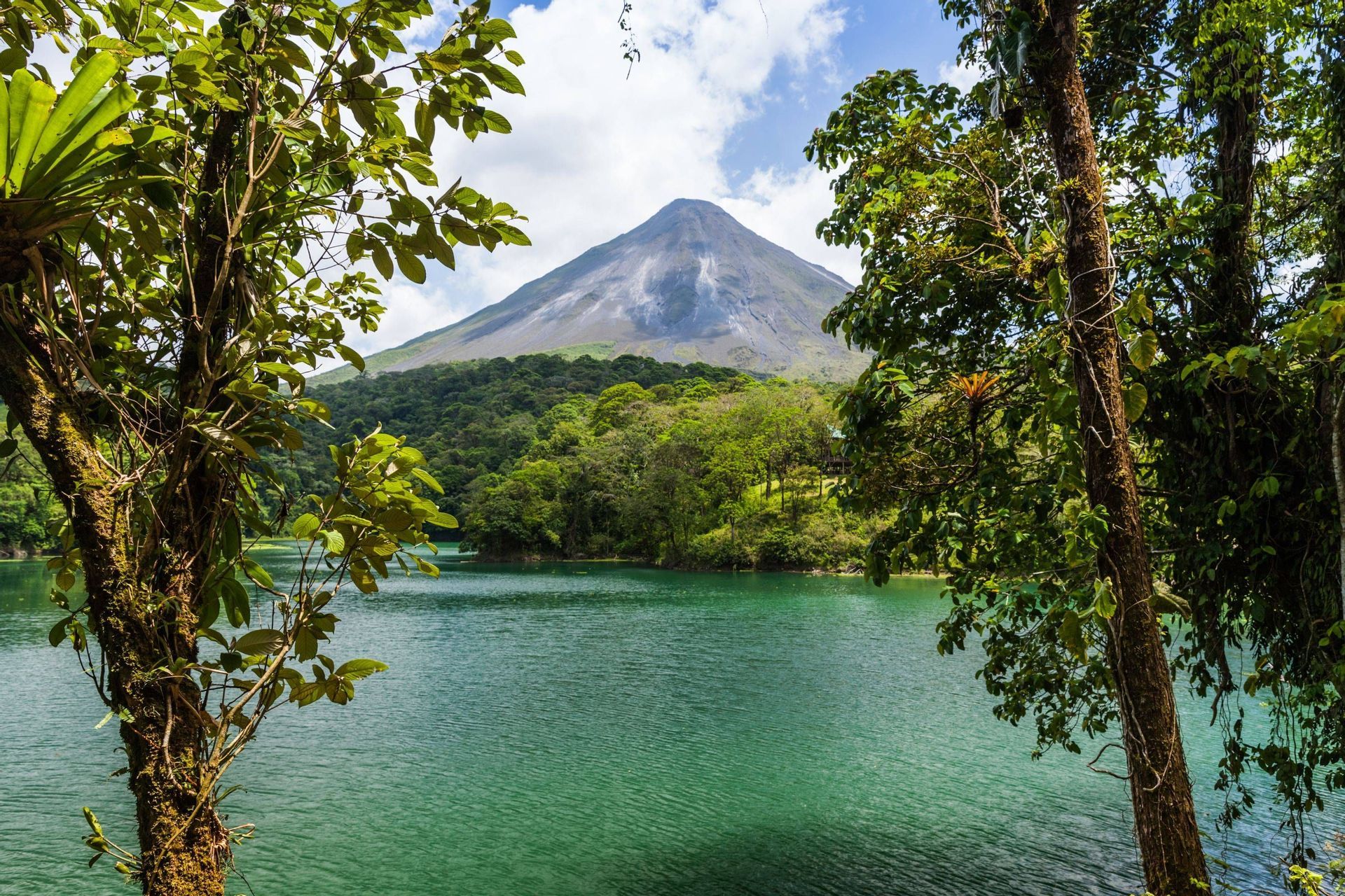 Un grande vulcano si erge sopra una fitta foresta e un lago verde, incorniciato da alberi frondosi sotto un cielo parzialmente nuvoloso.