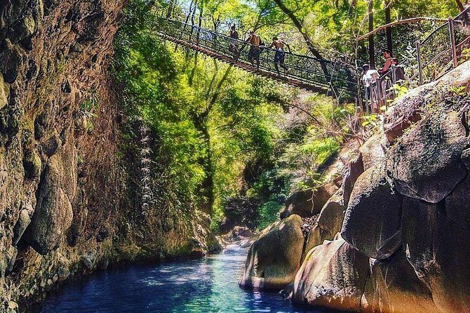 Un viaggio di gruppo WeRoad attraversa un ponte sospeso su un fiume turchese che scorre attraverso un canyon roccioso e alberato.