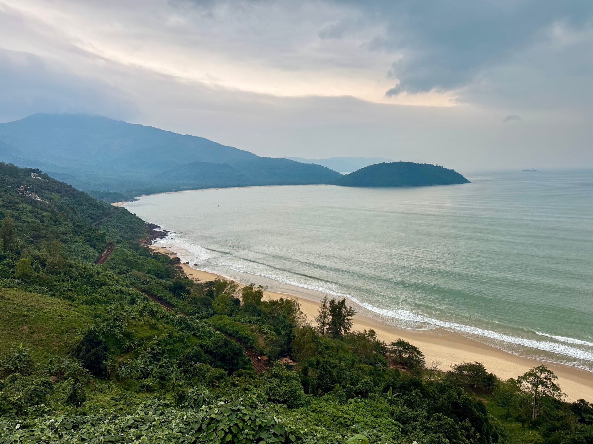 Una vista aérea de una costa tropical con una bahía de arena, exuberantes colinas verdes y montañas distantes bajo un cielo nublado.