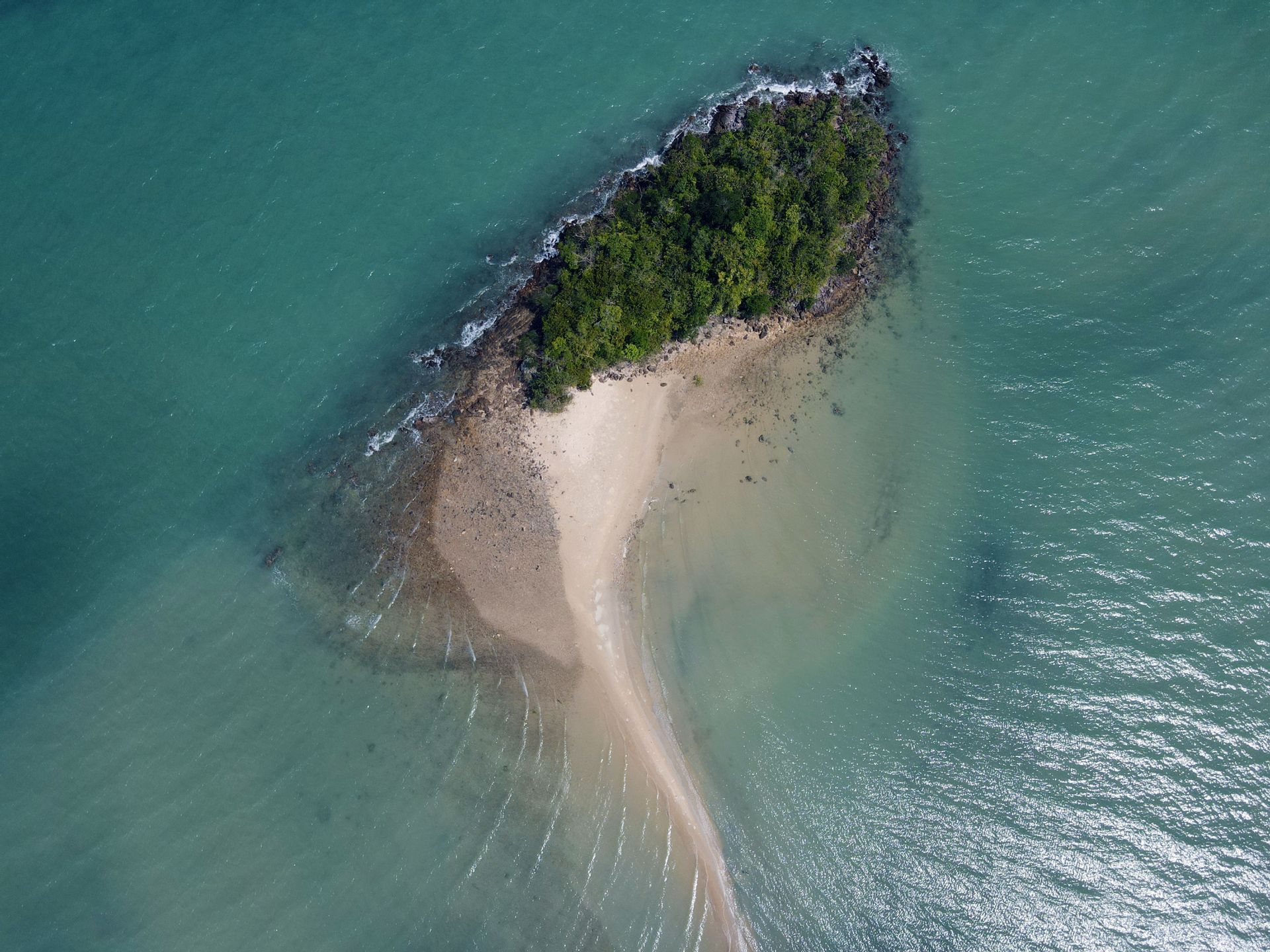 Una vista aérea de una pequeña isla cubierta de árboles con un banco de arena que se extiende hacia el agua tranquila de color turquesa.