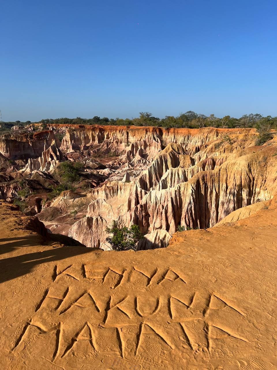 Le parole 'Kenya Hakuna Matata' incise nella sabbia arancione sul bordo di una scogliera, con vista su un vasto canyon multicolore.