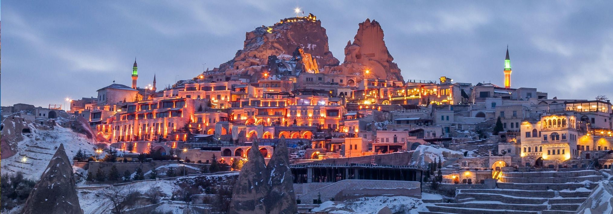 Une vue panoramique d'une ville enneigée construite à flanc de colline rocheuse, illuminée par des lumières chaleureuses au crépuscule.