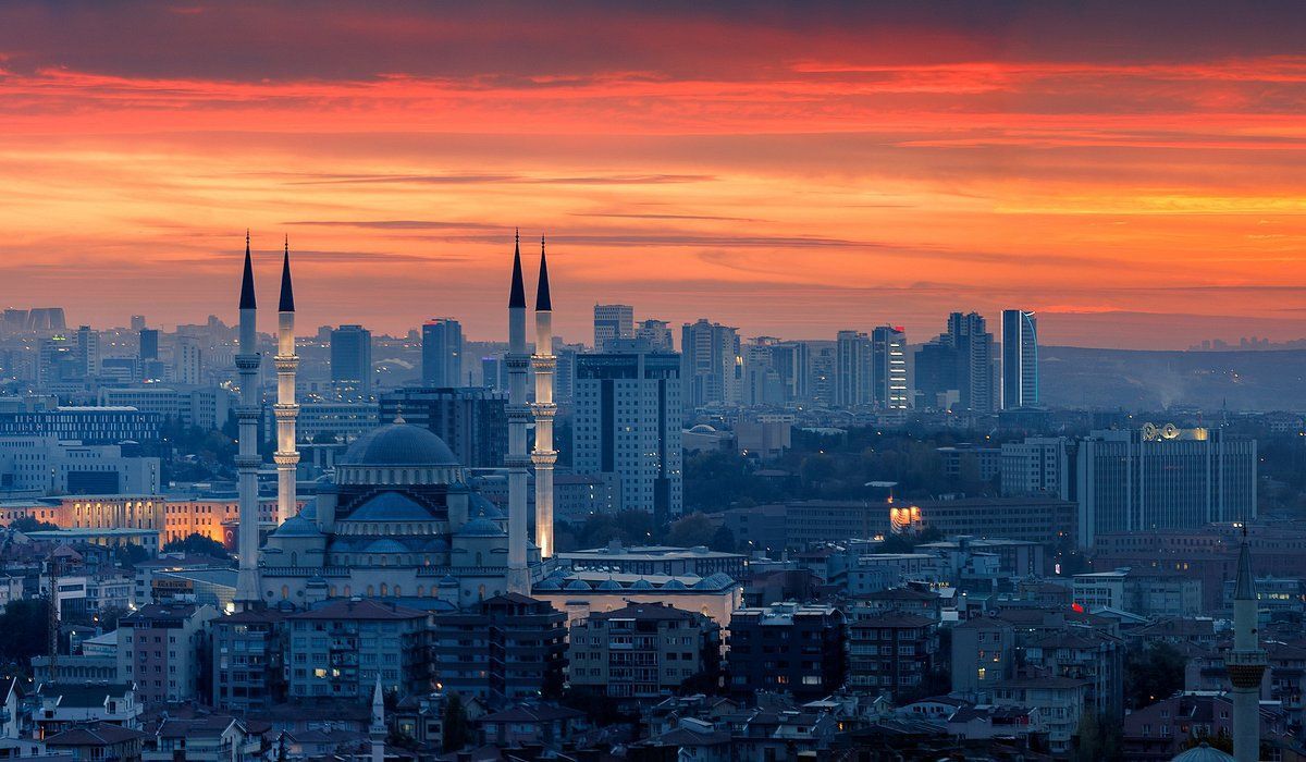 Une mosquée illuminée avec de hauts minarets se dresse dans un paysage urbain sur fond de coucher de soleil vibrant orange et rouge.