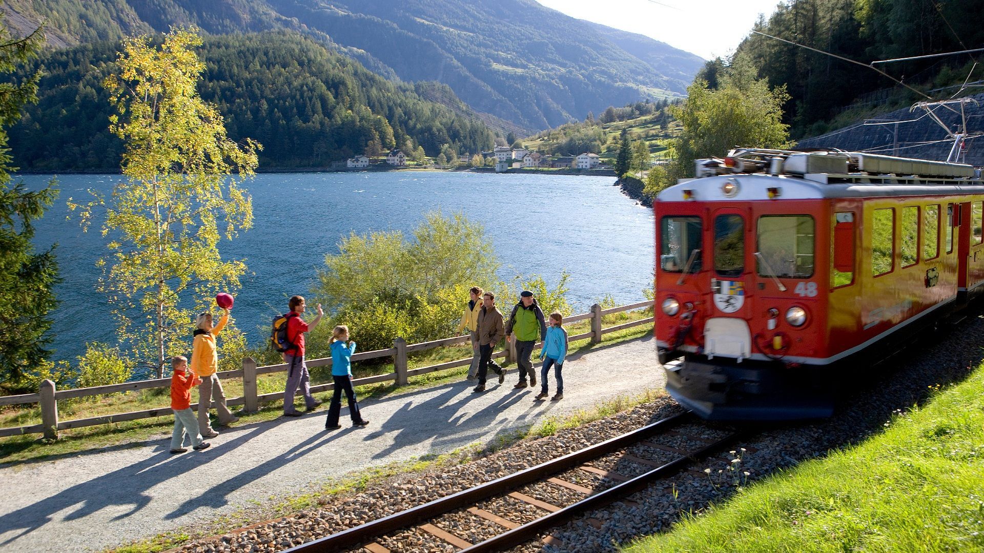 Un treno rosso corre su un binario accanto a un lago di montagna, mentre un gruppo di persone su un sentiero lo saluta.