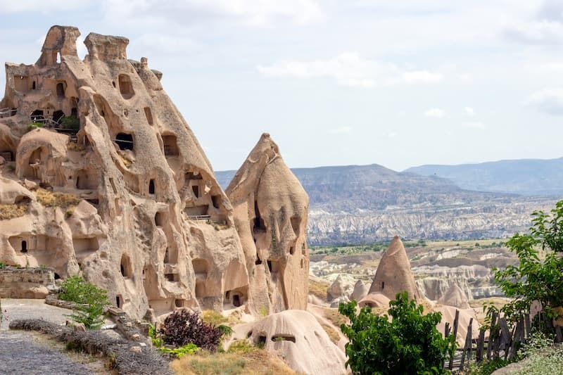Grandes formations rocheuses avec d'anciennes habitations troglodytiques sculptées, surplombant une vaste vallée avec des collines lointaines.