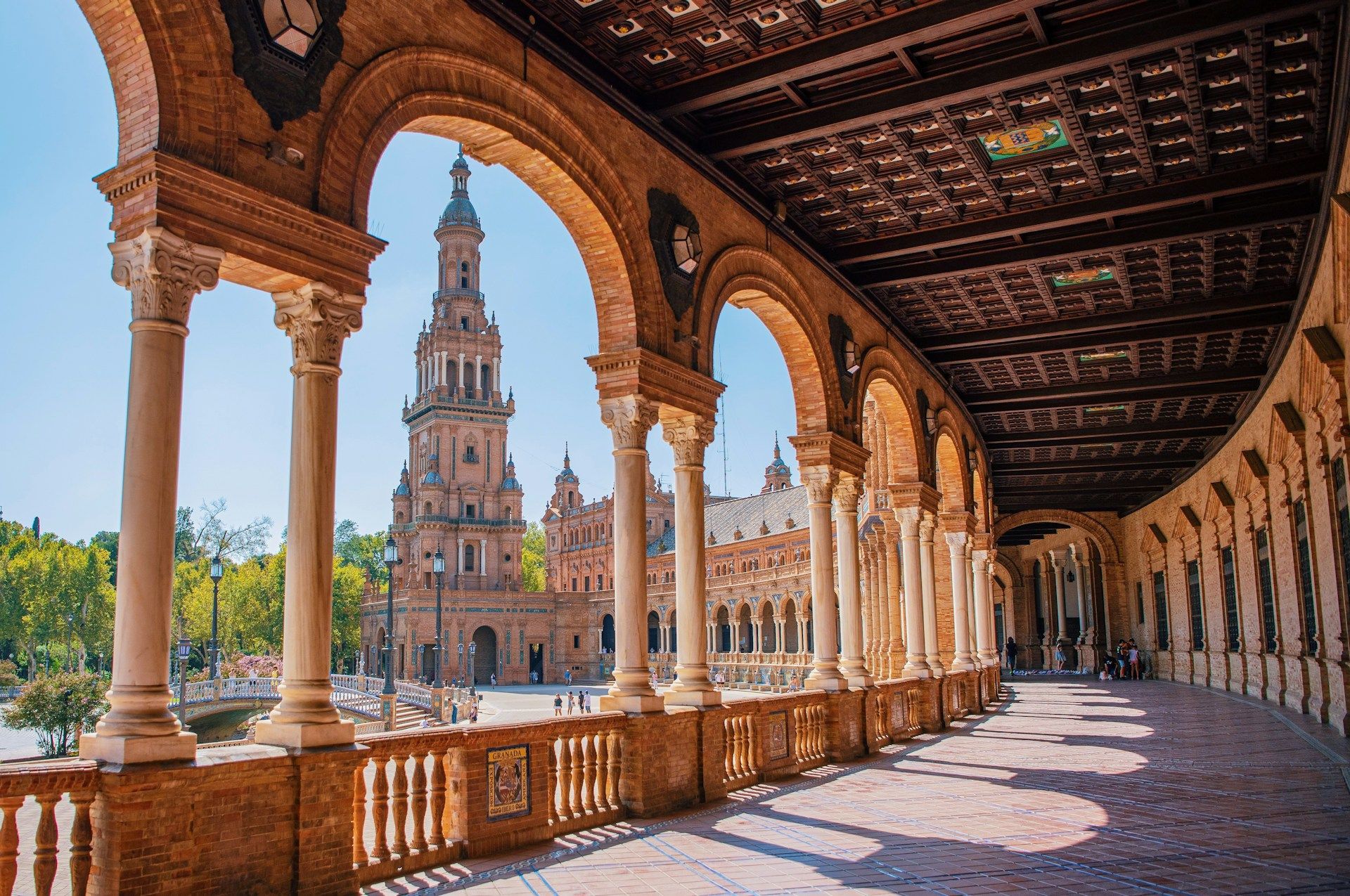 Una vista da una galleria curva con archi e colonne in mattoni che si affaccia su una piazza soleggiata con una torre decorata.