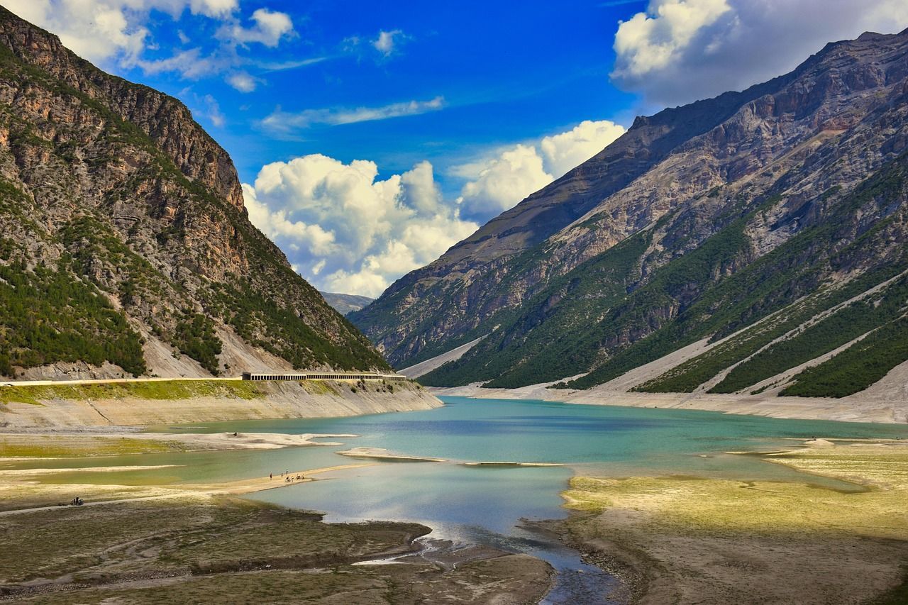Un lago turchese si snoda attraverso una profonda valle tra ripide montagne rocciose sotto un cielo blu con nuvole bianche.