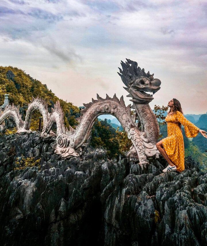 Una donna in abito giallo posa su una cima rocciosa accanto a una grande scultura di drago in pietra.