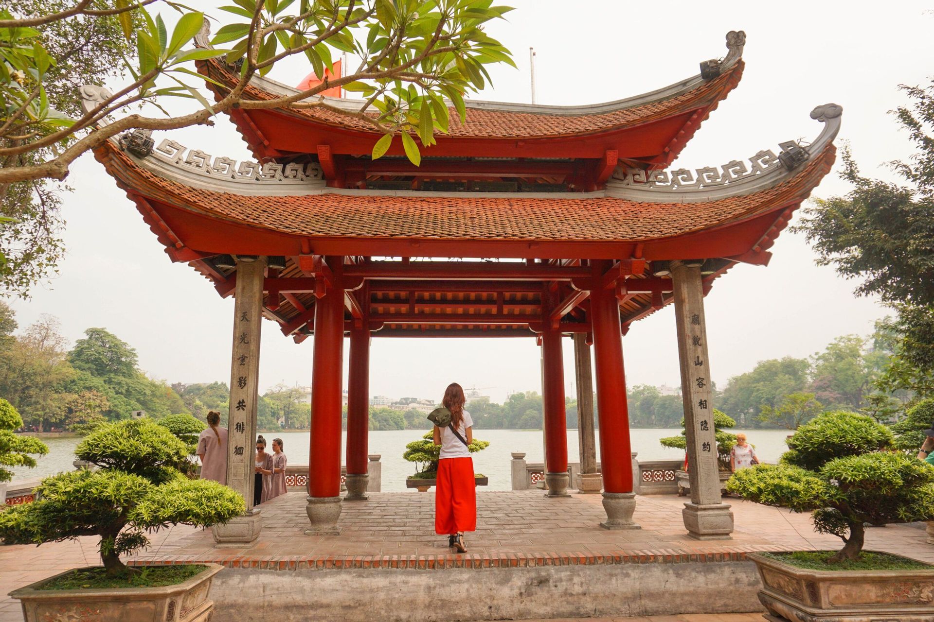 Una donna in pantaloni rossi si trova all'interno di una tradizionale pagoda rossa con tetto a tegole, guardando fuori su un lago con alberi e bonsai.