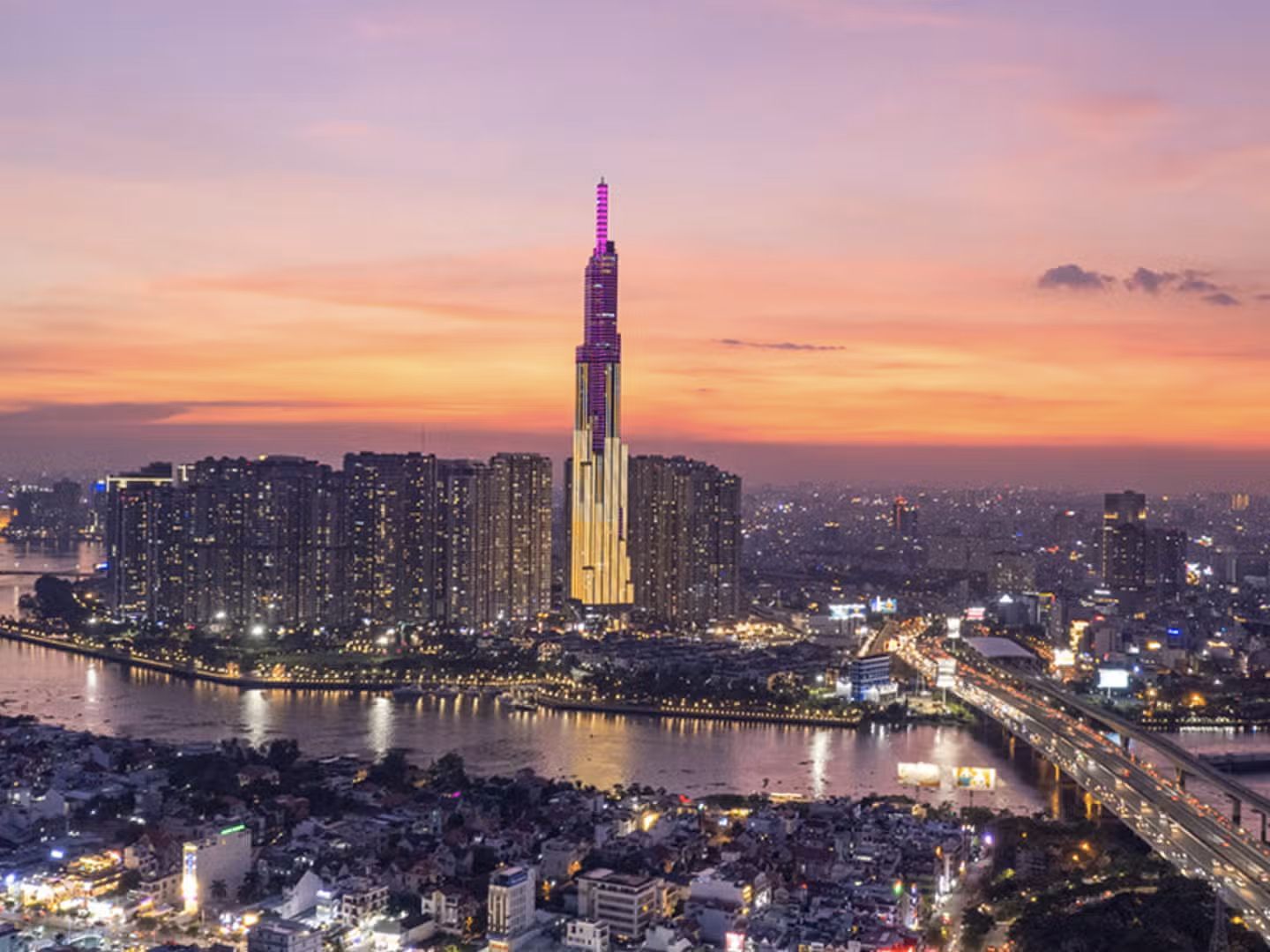 Un grattacielo illuminato con una guglia viola domina un denso skyline cittadino accanto a un ampio fiume al tramonto.