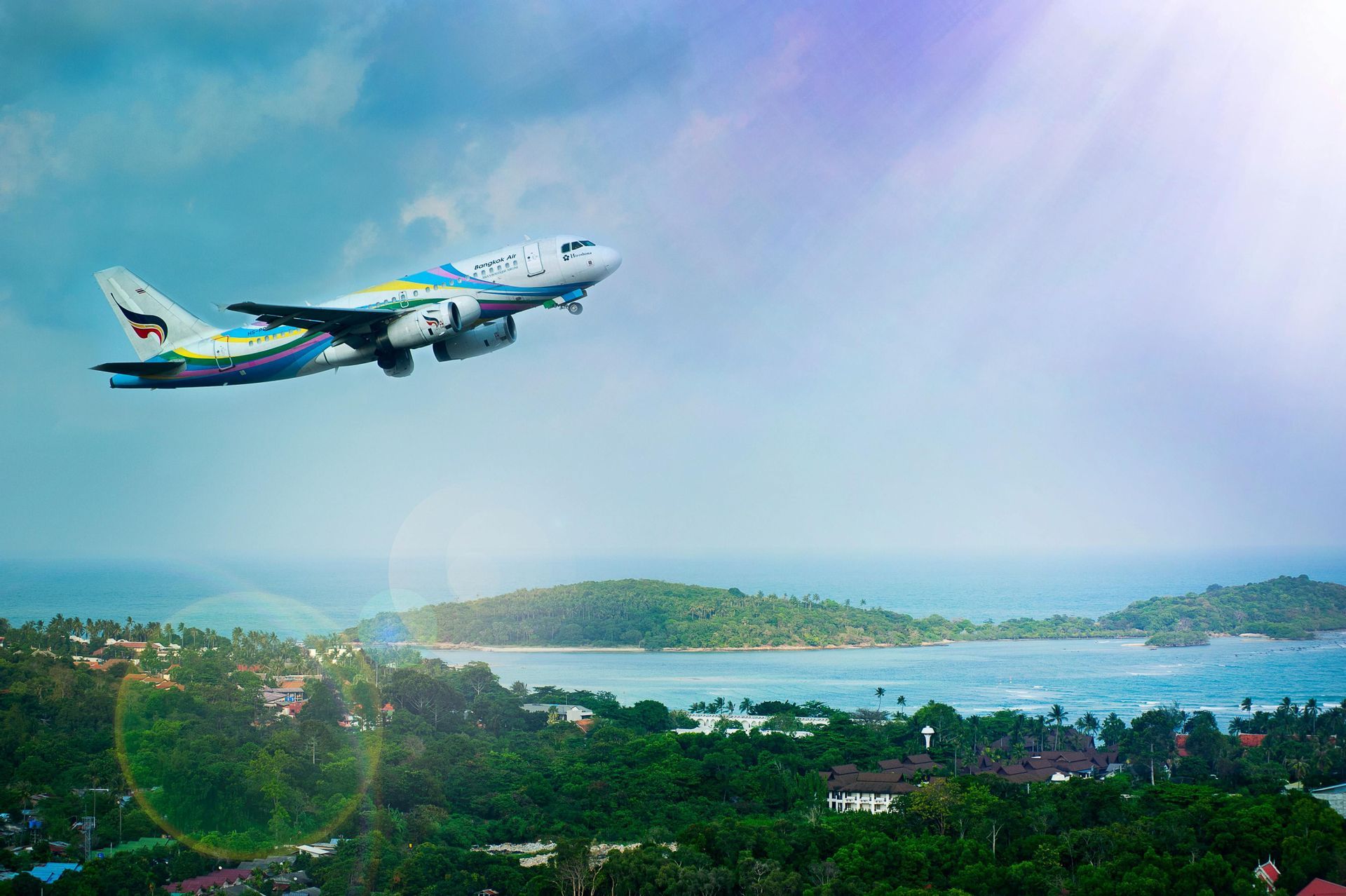 Un colorato aereo passeggeri vola basso su una lussureggiante costa tropicale con verdi colline e piccole isole nel mare.