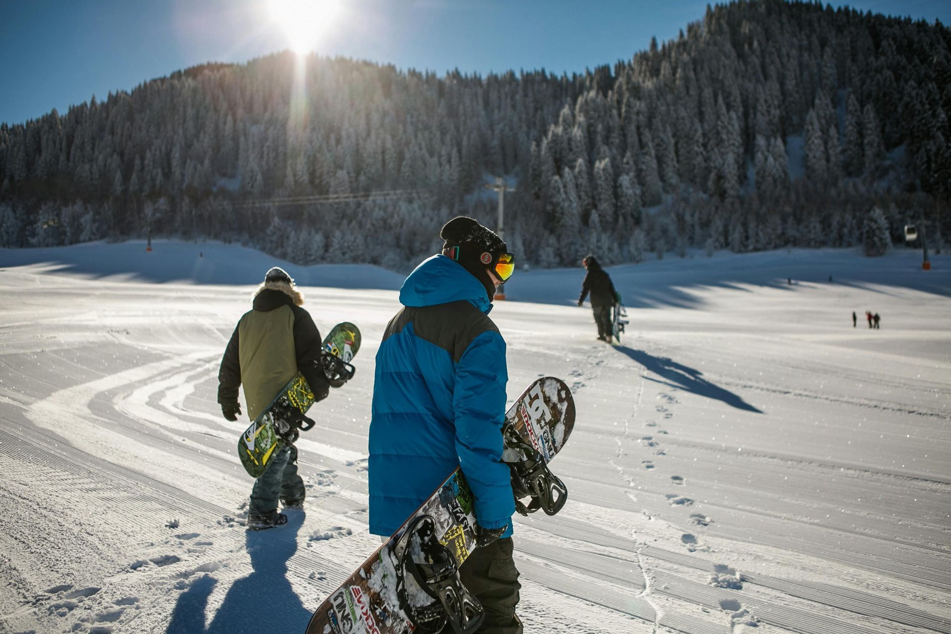 Un viaggio di gruppo WeRoad con tre persone che, portando gli snowboard, camminano su un pendio montano soleggiato e innevato.