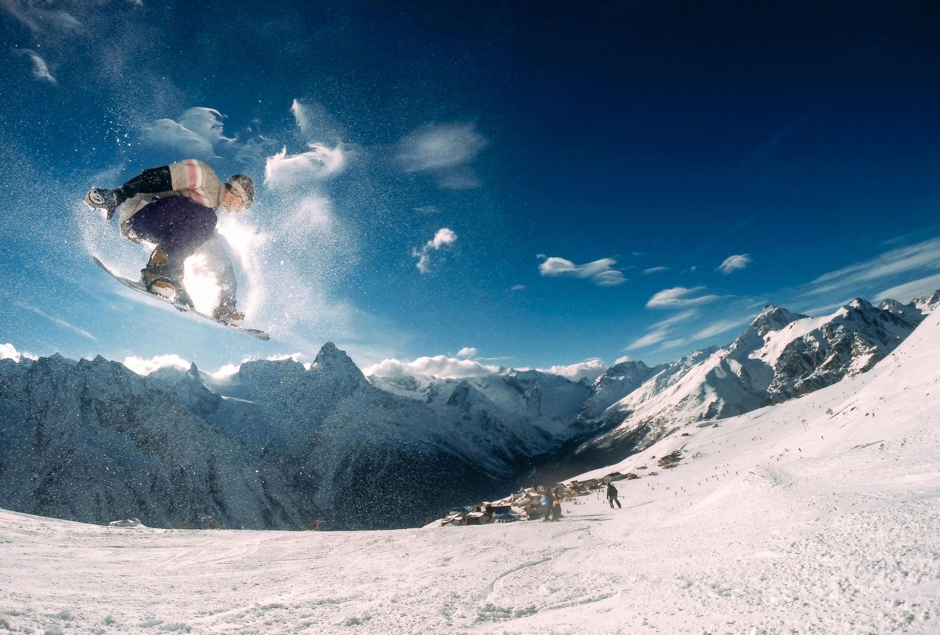 Uno snowboarder salta in aria sopra una catena montuosa innevata sotto un cielo azzurro chiaro.
