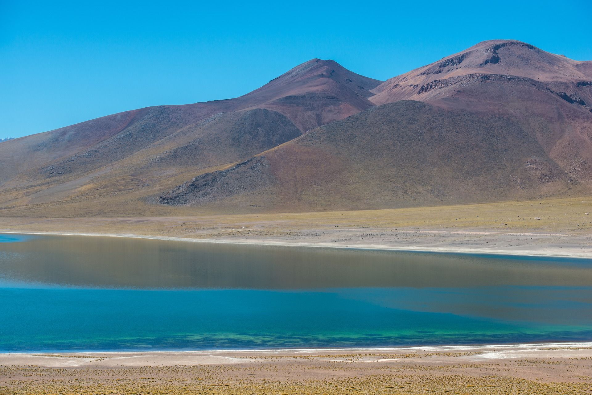 Un lago calmo e turchese si trova alla base di montagne aride e rossastre sotto un cielo azzurro e limpido.