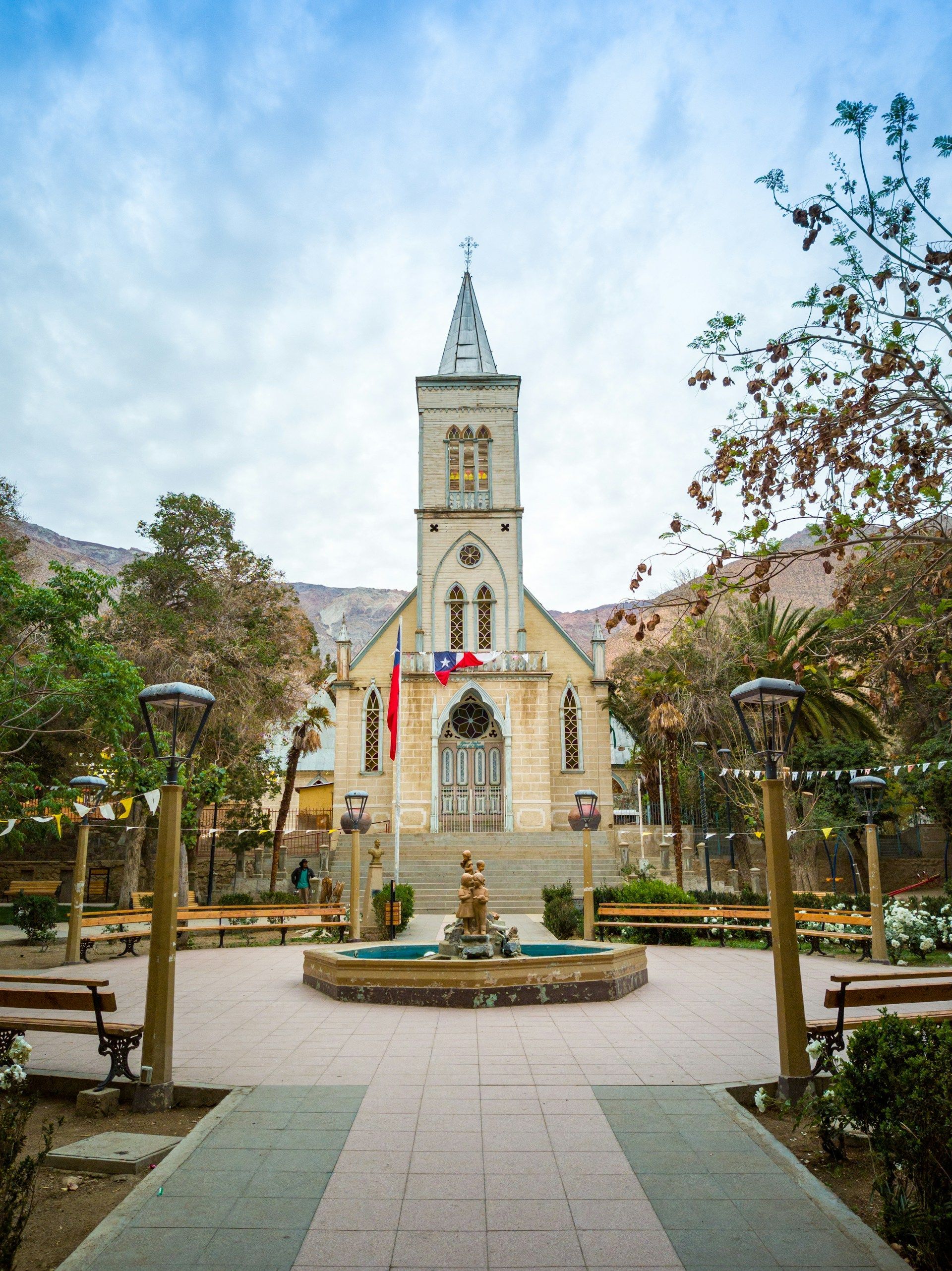 Una chiesa in pietra con un alto campanile e due bandiere si erge in una piazza con una fontana, circondata da alberi e montagne.