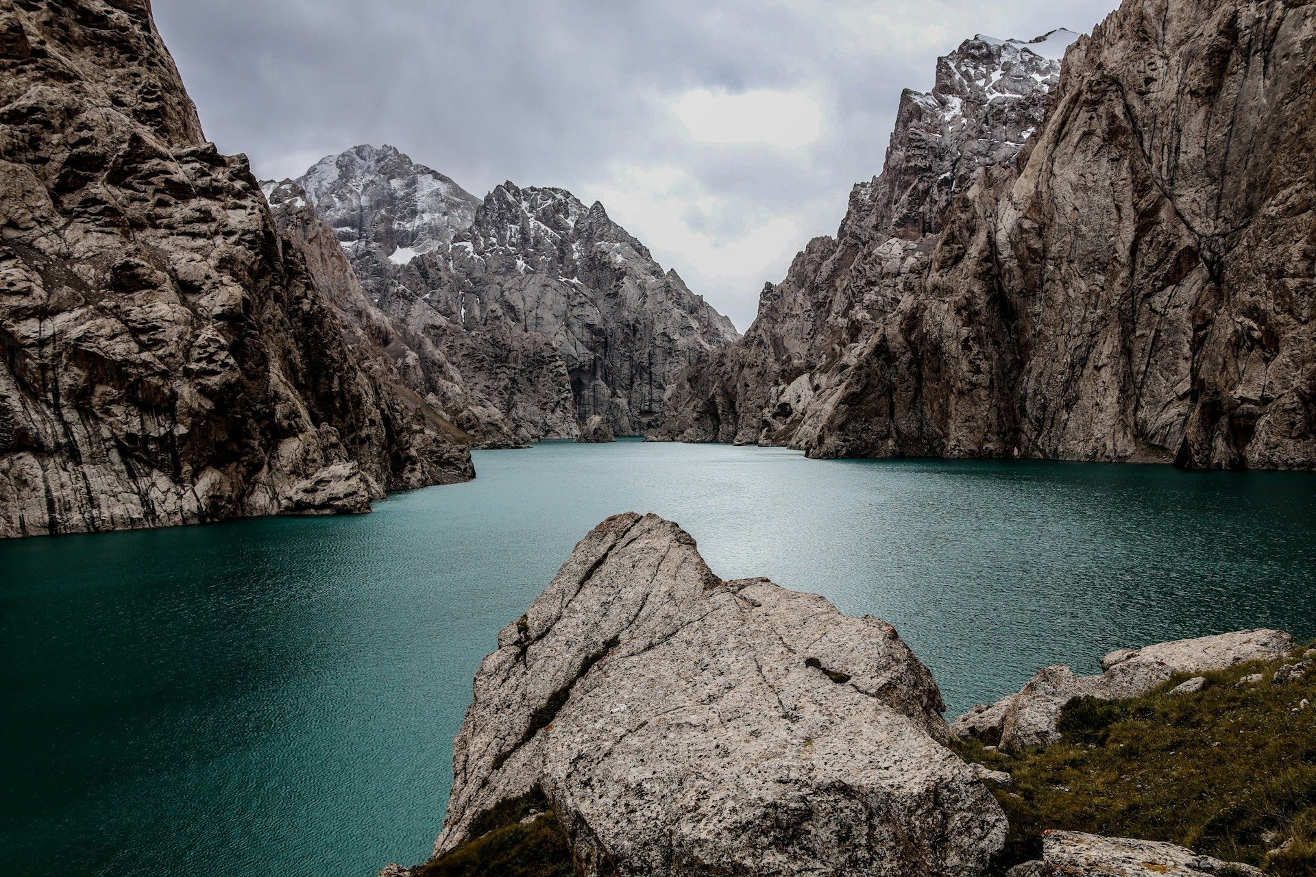 Un lago turchese si snoda attraverso un aspro canyon di montagna con cime innevate sotto un cielo nuvoloso.