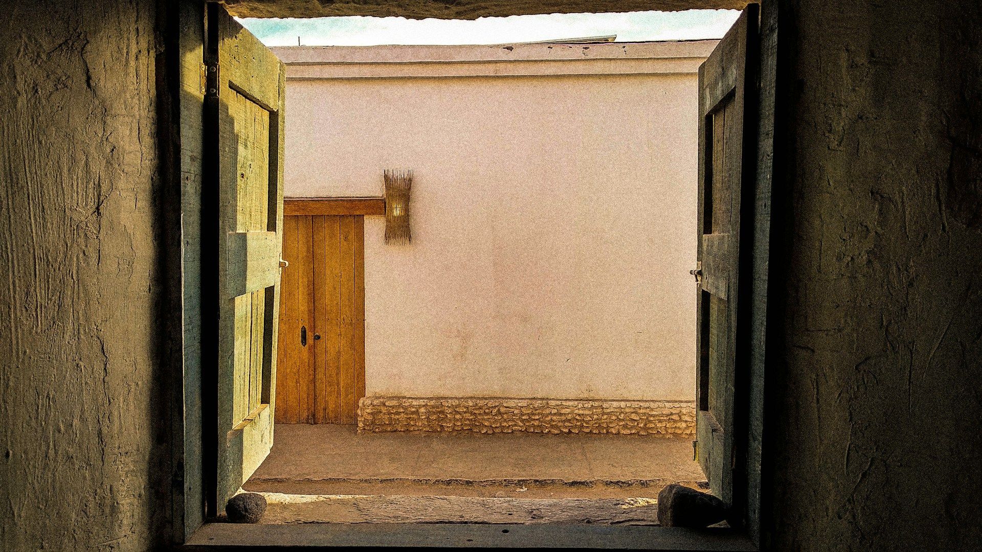 Vista da un interno buio attraverso porte di legno aperte su un cortile soleggiato con un muro imbiancato e un'altra porta.