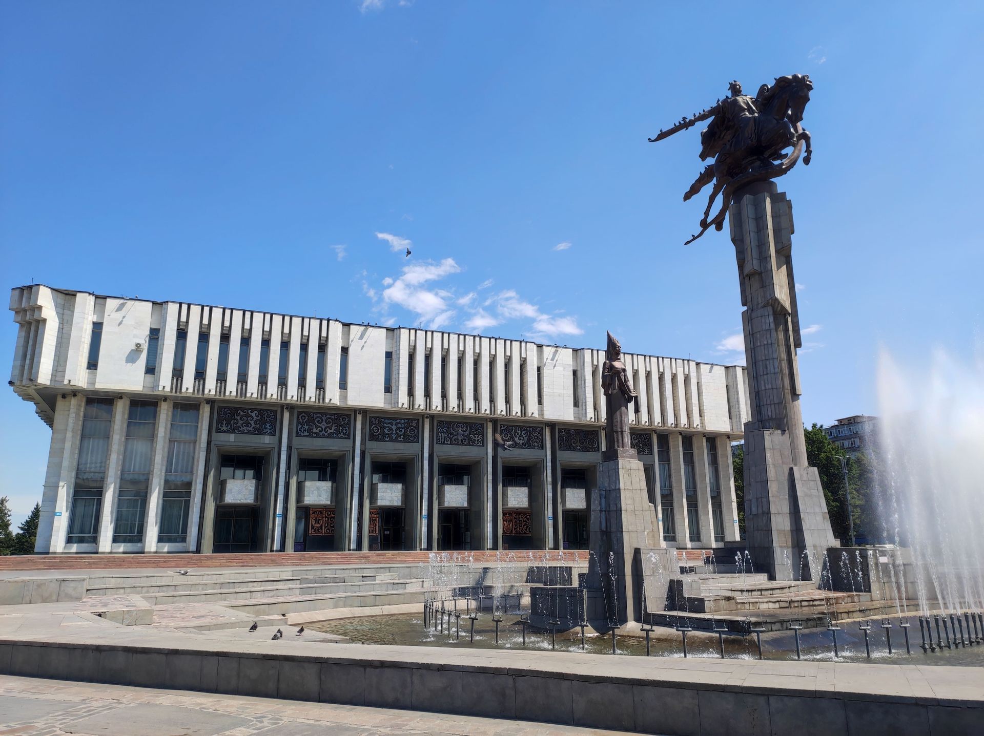 Una estatua ecuestre monumental y una fuente en una plaza frente a un gran edificio blanco bajo un cielo azul claro.
