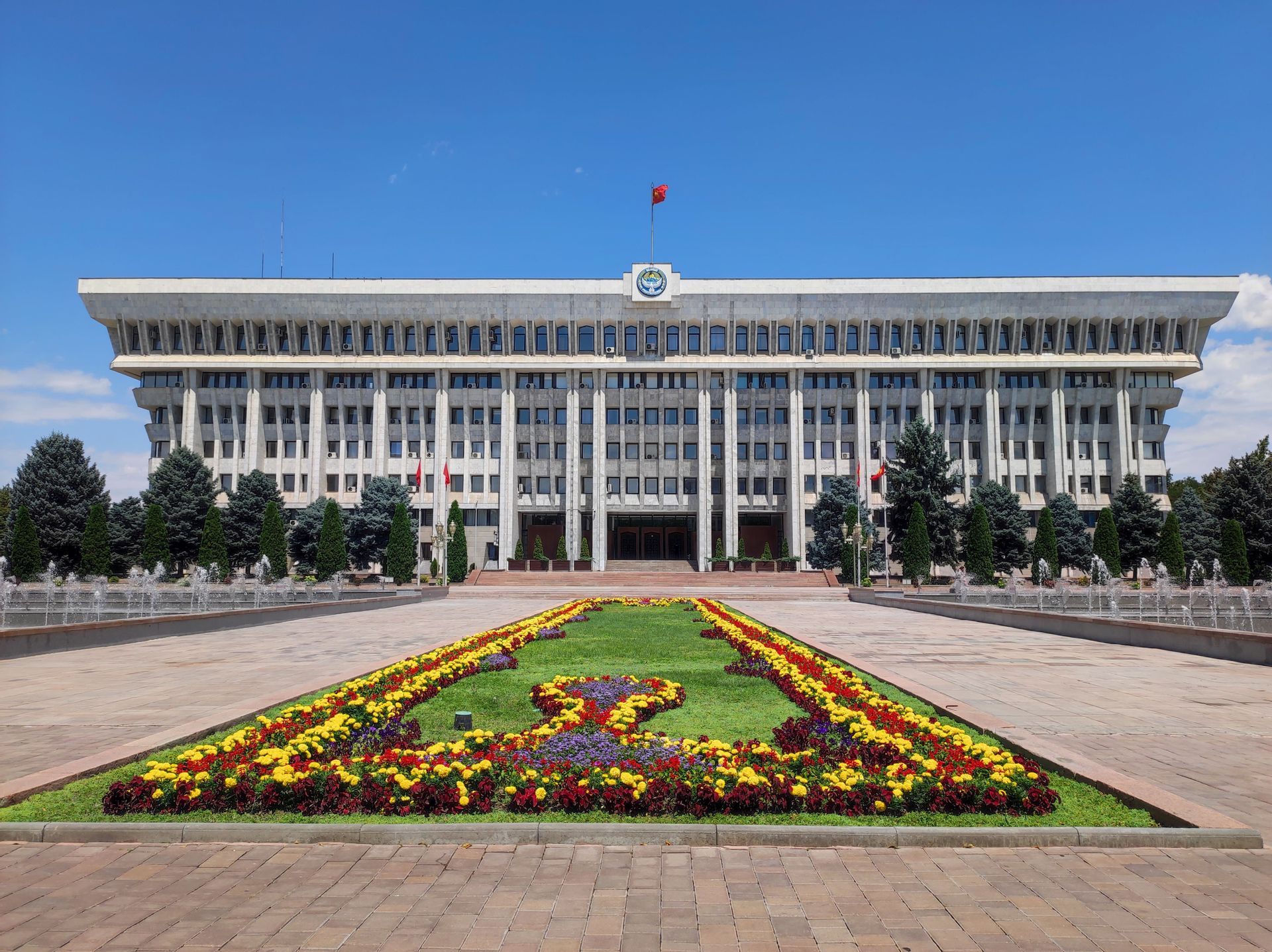 A large government building with a flag on top, viewed from across a plaza with fountains and colorful flowerbeds.