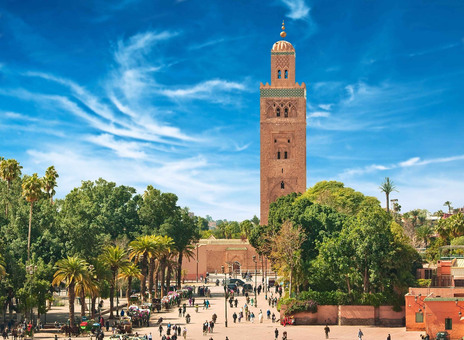 Un alto minareto color sabbia si erge su una vivace piazza cittadina piena di persone e palme sotto un cielo blu.
