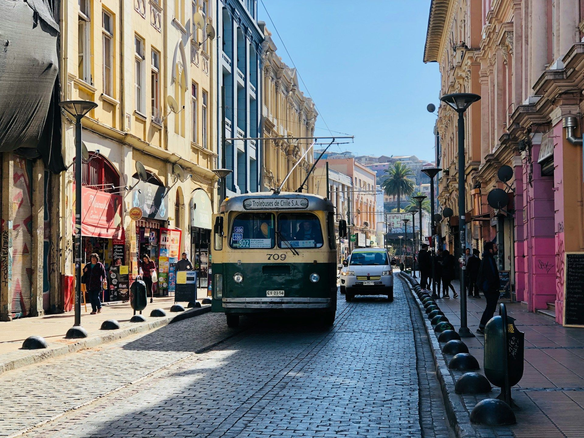 Un filobus vintage verde e crema percorre una strada acciottolata tra edifici colorati in una giornata di sole.